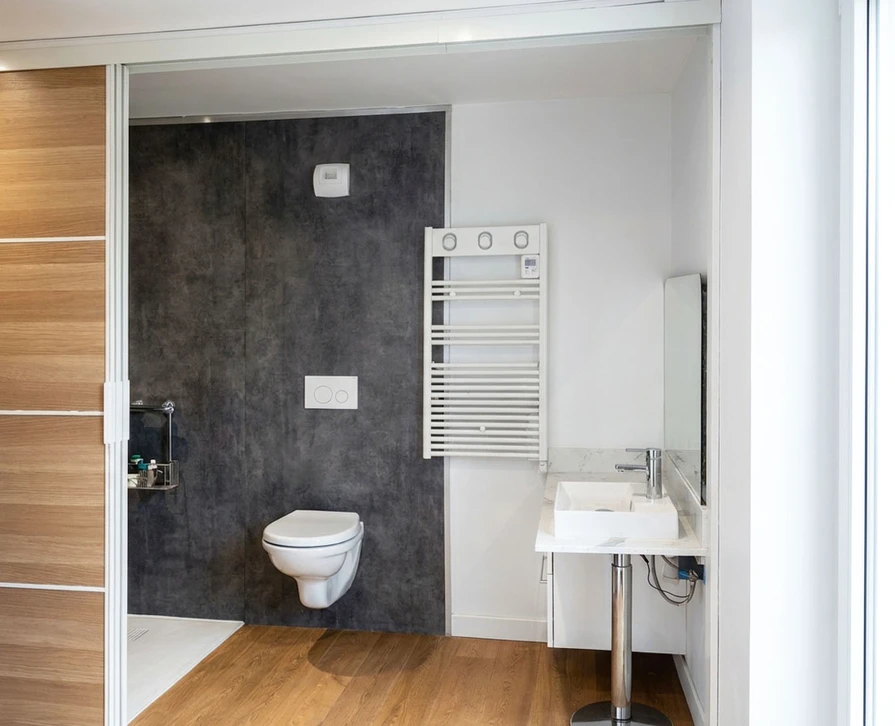 Un électricien professionnel en tenue de travail effectuant des travaux de mise aux normes électrique dans un salon d'une maison en rénovation. En arrière-plan, visible mais légèrement flou, on distingue d'autres éléments de chantier suggérant des travaux multi-domaines : des tuyaux de ventilation, des panneaux d'isolation thermique contre un mur, et une partie de porte entrouverte donnant accès à une salle de bain. La scène doit transmettre l'idée d'un professionnel gérant l'ensemble d'un projet de rénovation globale. Lumière naturelle ou semi-professionnelle, ambiance de chantier réel.